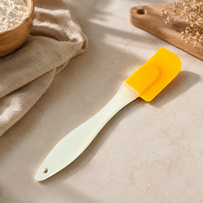 A yellow silicone spatula with a light cream handle rests on a marble counter next to a bowl of flour and a beige cloth.