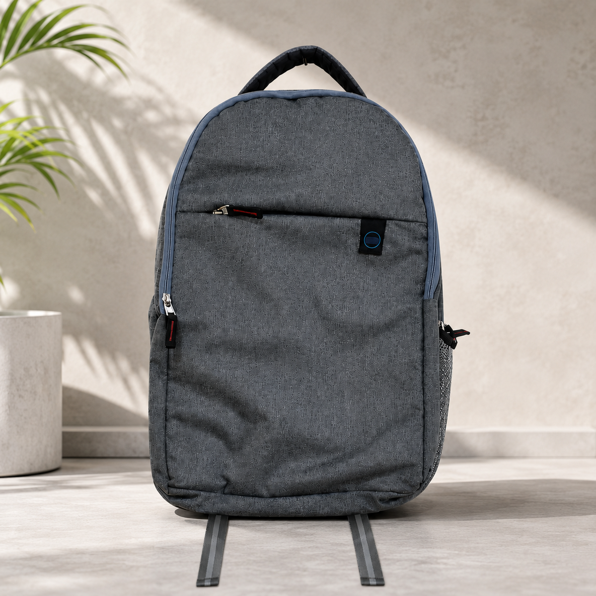 A grey backpack with a front zipper pocket and blue trim stands on a light grey surface next to a plant.