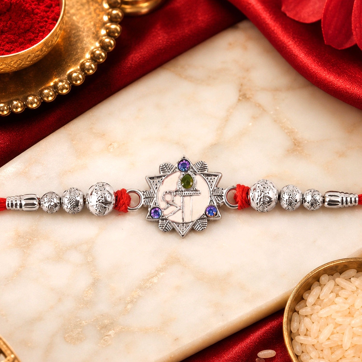 A silver Swastik charm with a green and purple gem center is flanked by silver beads on a red thread, resting on a marble