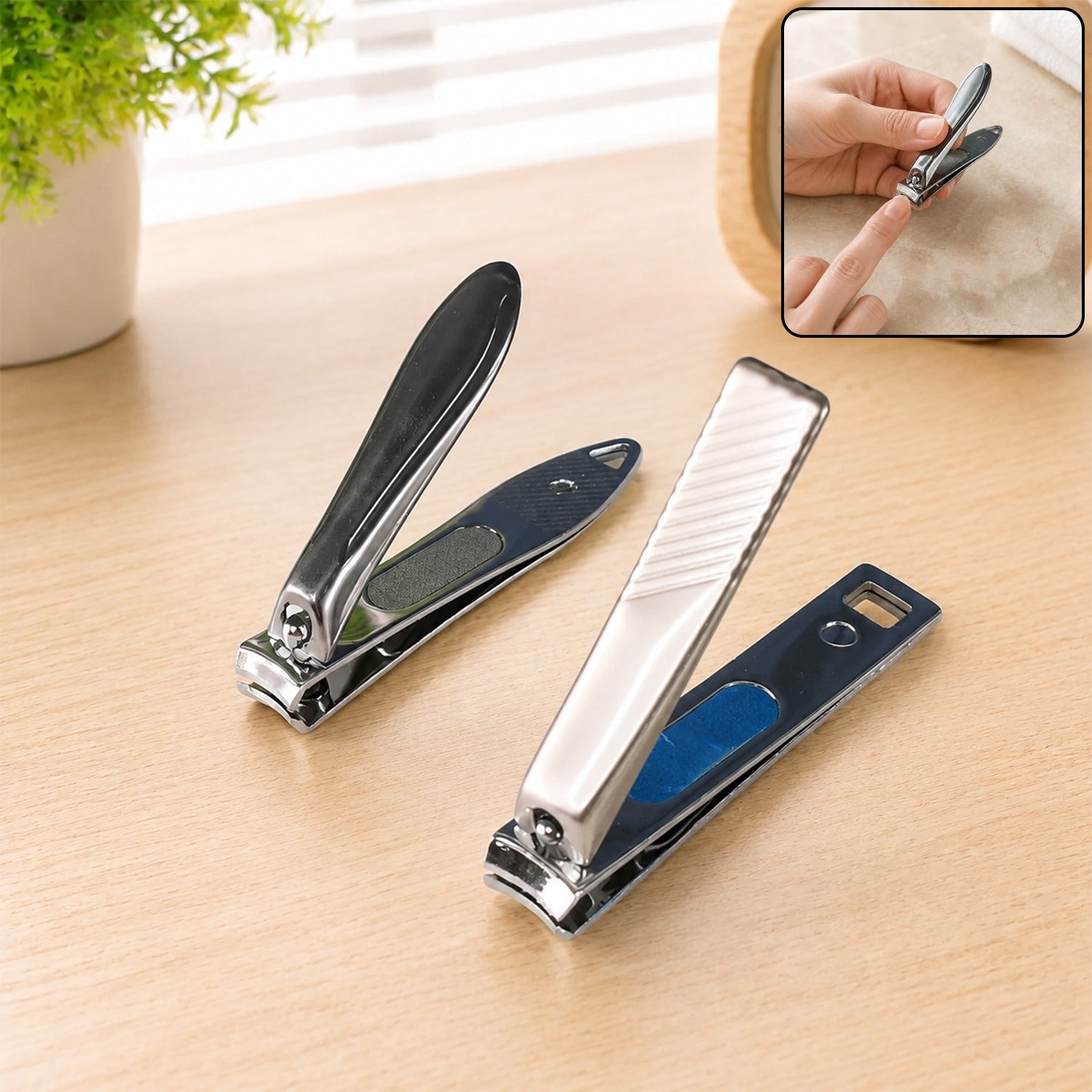 Two stainless steel nail clippers, one dark grey and one silver, on a wooden table with a plant.