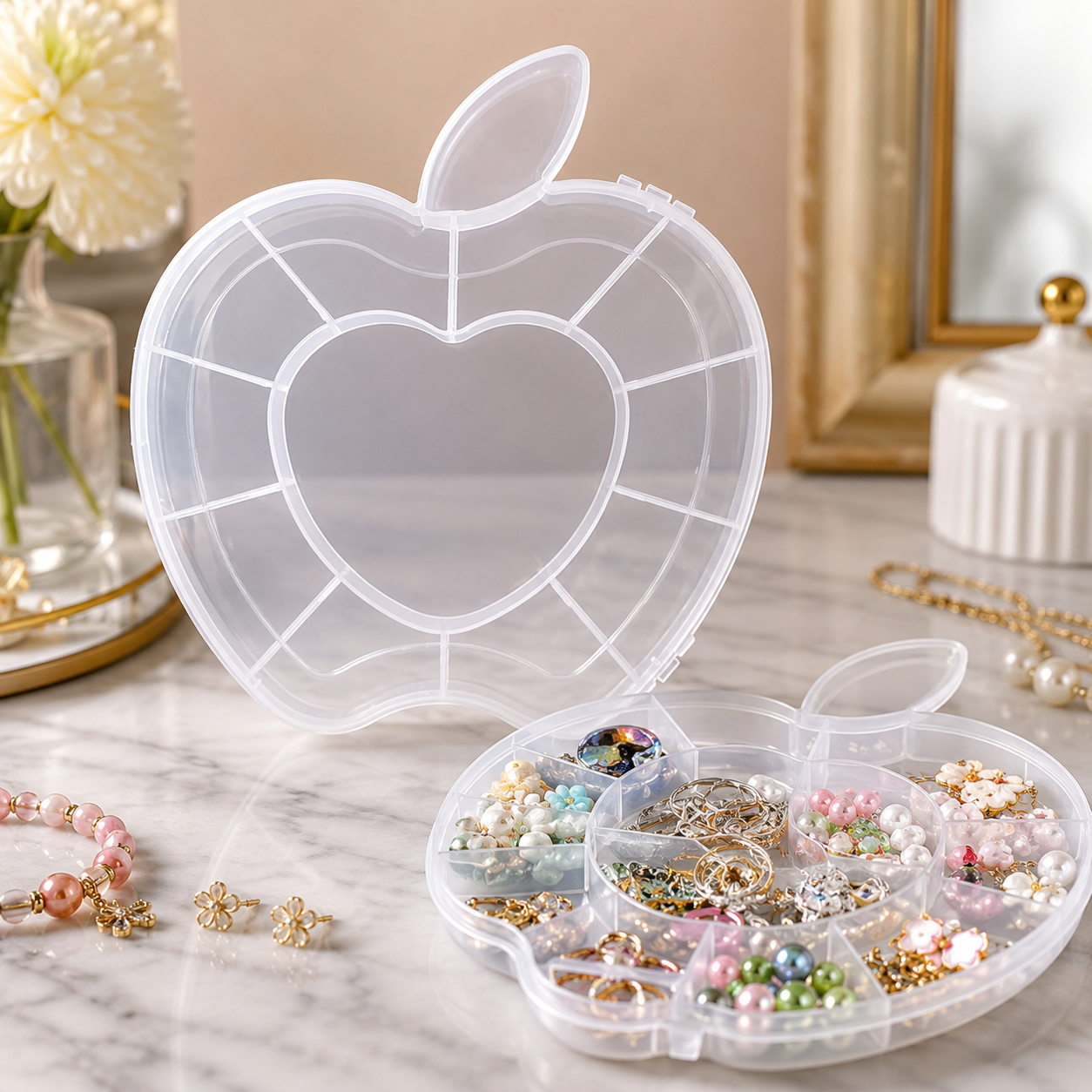 A transparent apple-shaped organizer box with multiple compartments, filled with various colorful jewelry, sits on a marble