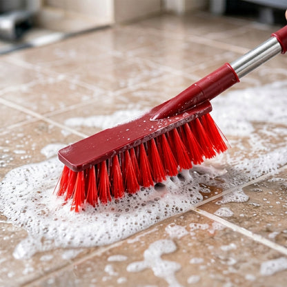A red and silver brush with red bristles is scrubbing a tiled floor covered in white soap suds.