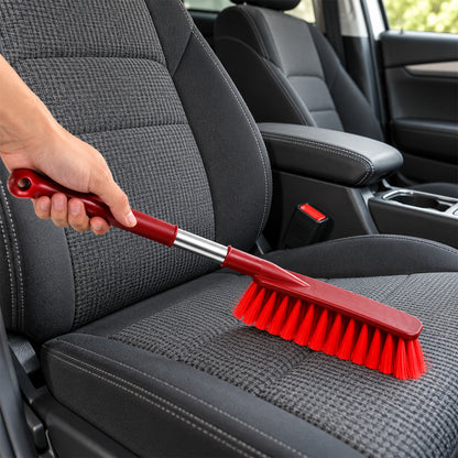 A hand uses a red and silver brush to clean the gray fabric upholstery of a car seat.