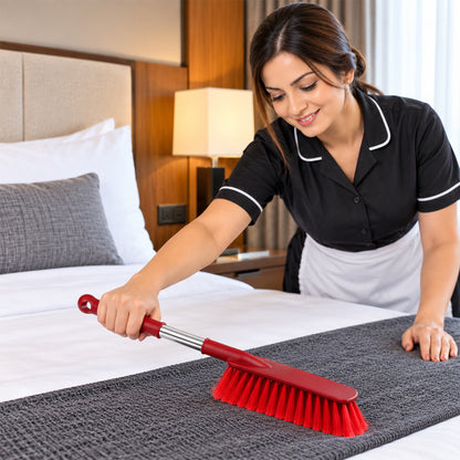 A smiling woman in a black and white uniform uses a red and silver brush to clean a gray bed runner.