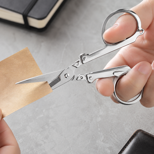 A hand holds silver folding scissors, cutting a piece of brown paper against a grey textured background.