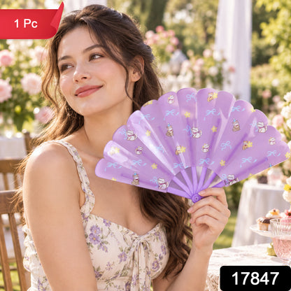 A woman smiles while holding a purple fan with cat and bow designs in a sunny outdoor garden.