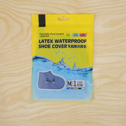A yellow and clear plastic package for latex waterproof shoe covers, with text and a cartoon shoe, rests on a light wood