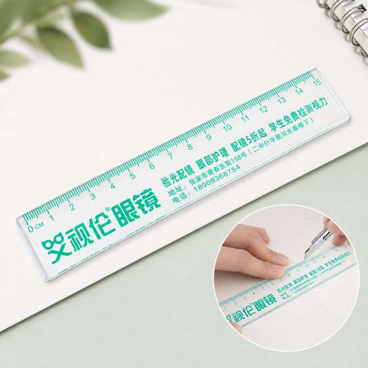 A transparent plastic 15cm ruler with green text and markings rests on a white notebook, next to a small inset image of a