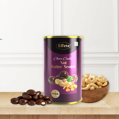 A purple and gold tin of chocolate-coated nuts sits on a wooden surface next to a bowl of cashews and a pile of chocolate