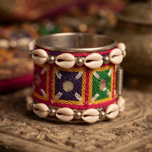 A vibrant cuff bracelet with mirror work, colorful embroidery, and cowrie shells, resting on a carved wooden surface.