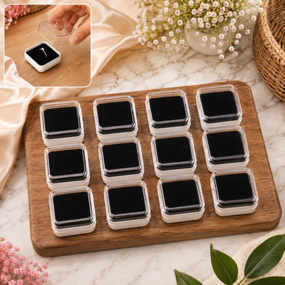 A hand opening a clear-lidded white box with a ring inside, surrounded by 11 other empty boxes on a wooden board.