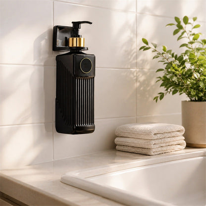 A black wall-mounted soap dispenser with a gold pump and accent, next to a potted plant and folded towels.
