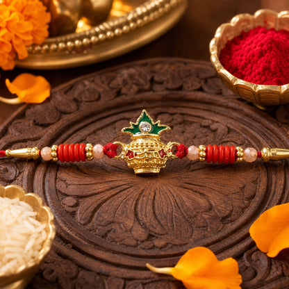 A gold kalash charm with green enamel and a clear gem sits on a red string with gold beads, on a carved wooden surface.