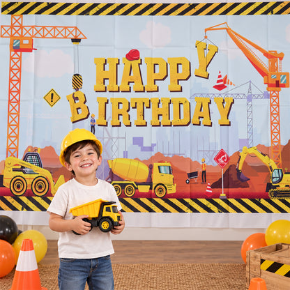 A young boy in a yellow hard hat smiles while holding a toy dump truck in front of a construction birthday banner.