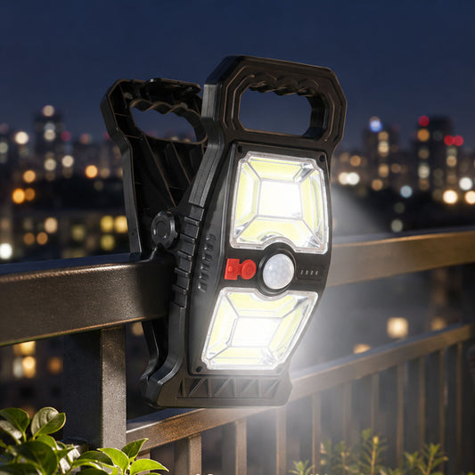 A black solar motion sensor clip light illuminates a dark city balcony railing at night, with distant buildings blurred.