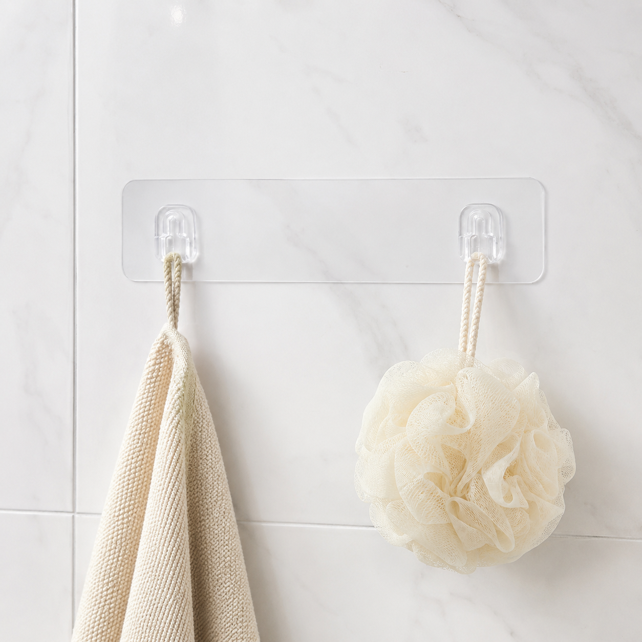 A clear adhesive wall hook strip with two hooks, holding a beige towel and a cream bath pouf against a white tiled wall.