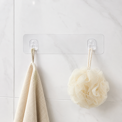 A clear adhesive wall hook strip with two hooks, holding a beige towel and a cream bath pouf against a white tiled wall.