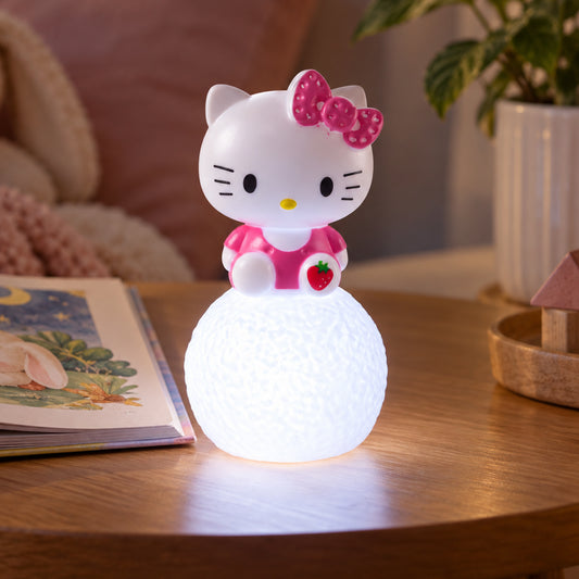 A Hello Kitty LED night light illuminates a wooden table next to a children's book and a potted plant.