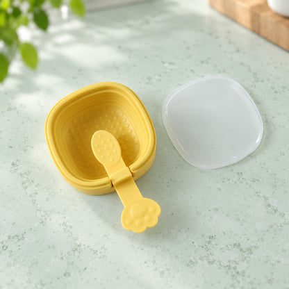 A yellow square silicone mold with a paw handle and a clear lid rests on a light green textured surface with a plant in the