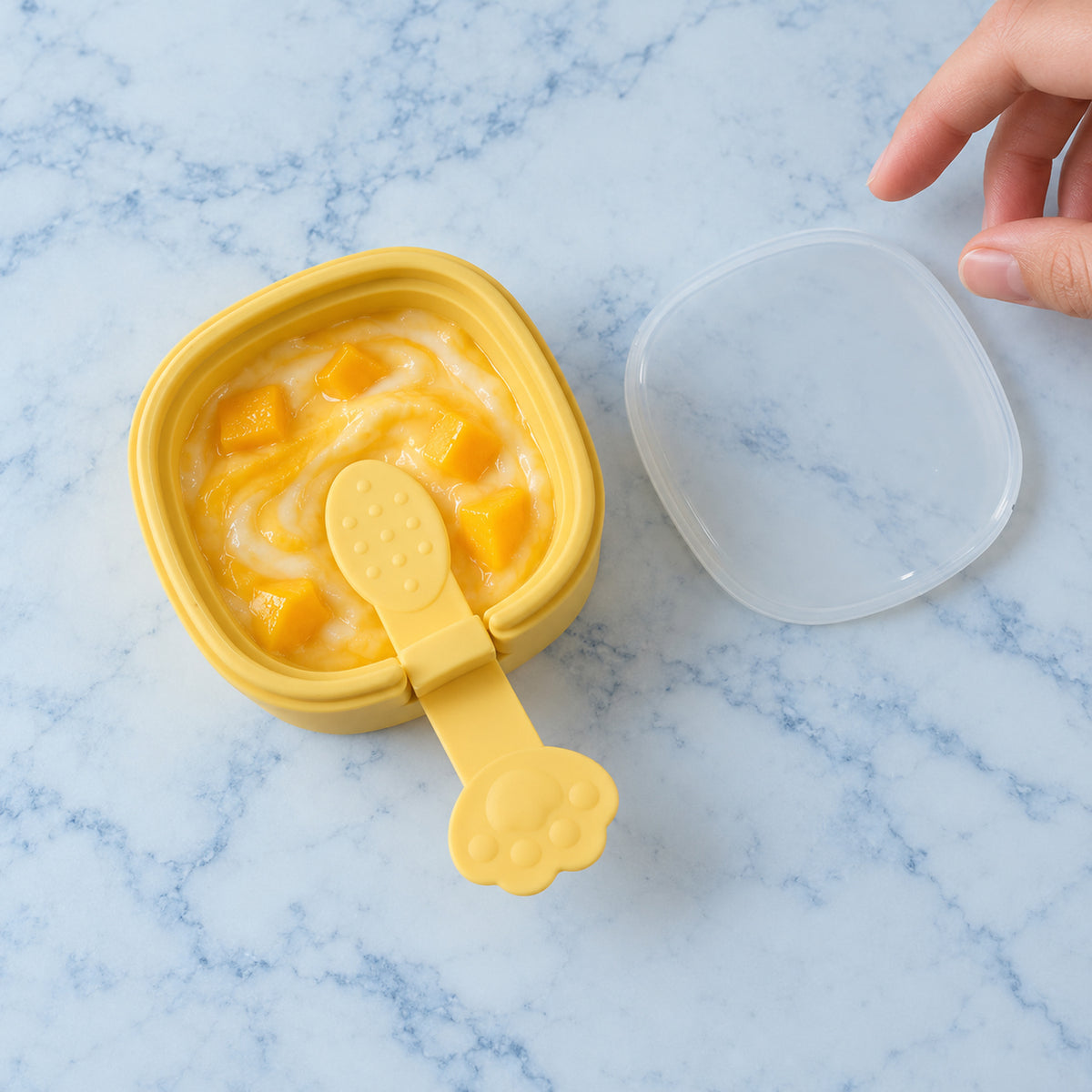 A hand reaches for a clear lid next to a yellow square mold filled with yellow mango and white yogurt mixture on a marble