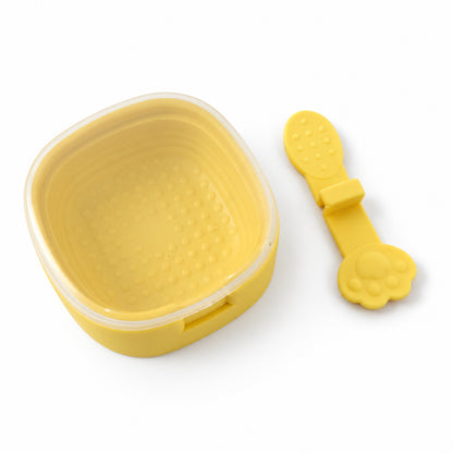 A yellow square silicone mold with a clear lid sits next to a detached yellow paw-shaped popsicle stick on a white surface.