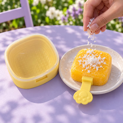 A hand sprinkles white coconut flakes onto a yellow square popsicle on a white plate, next to an open yellow mold.