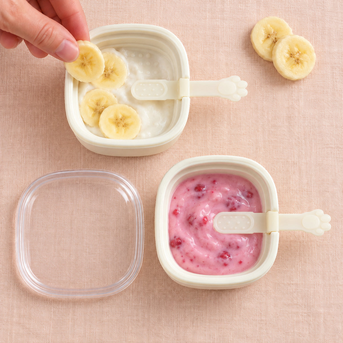 Two white square molds contain fruit purees, one with banana slices, the other with raspberries, on a pink surface.