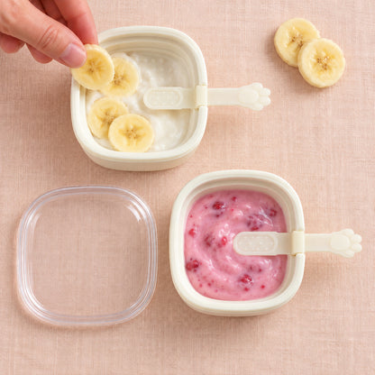 Two white square molds contain fruit purees, one with banana slices, the other with raspberries, on a pink surface.