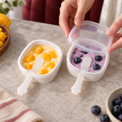 Two square molds with paw handles hold fruit ice creams, one yellow mango, one purple blueberry, on a speckled table.