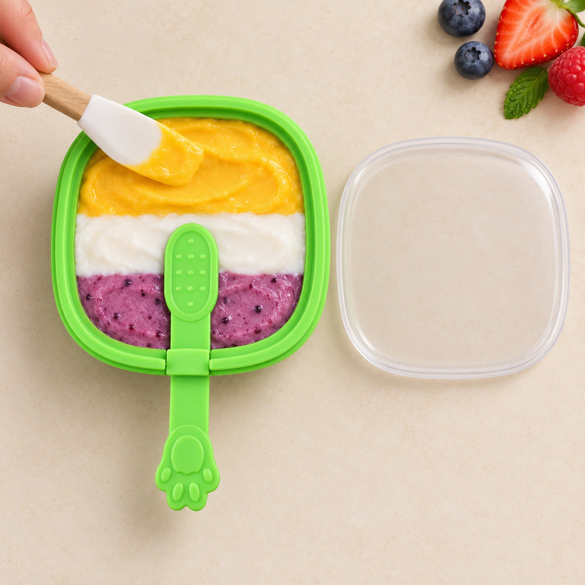 A hand uses a spatula to layer yellow, white, and purple purees into a green silicone mold with a paw handle.