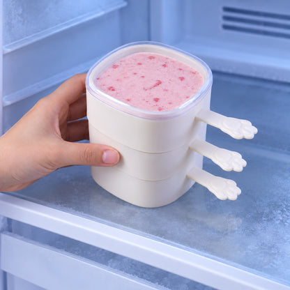 A hand placing a stack of three square molds, the top one filled with pink puree, into a frosty freezer.