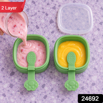 A hand pours pink liquid into a green square mold, beside another mold with yellow liquid, on a purple patterned surface.