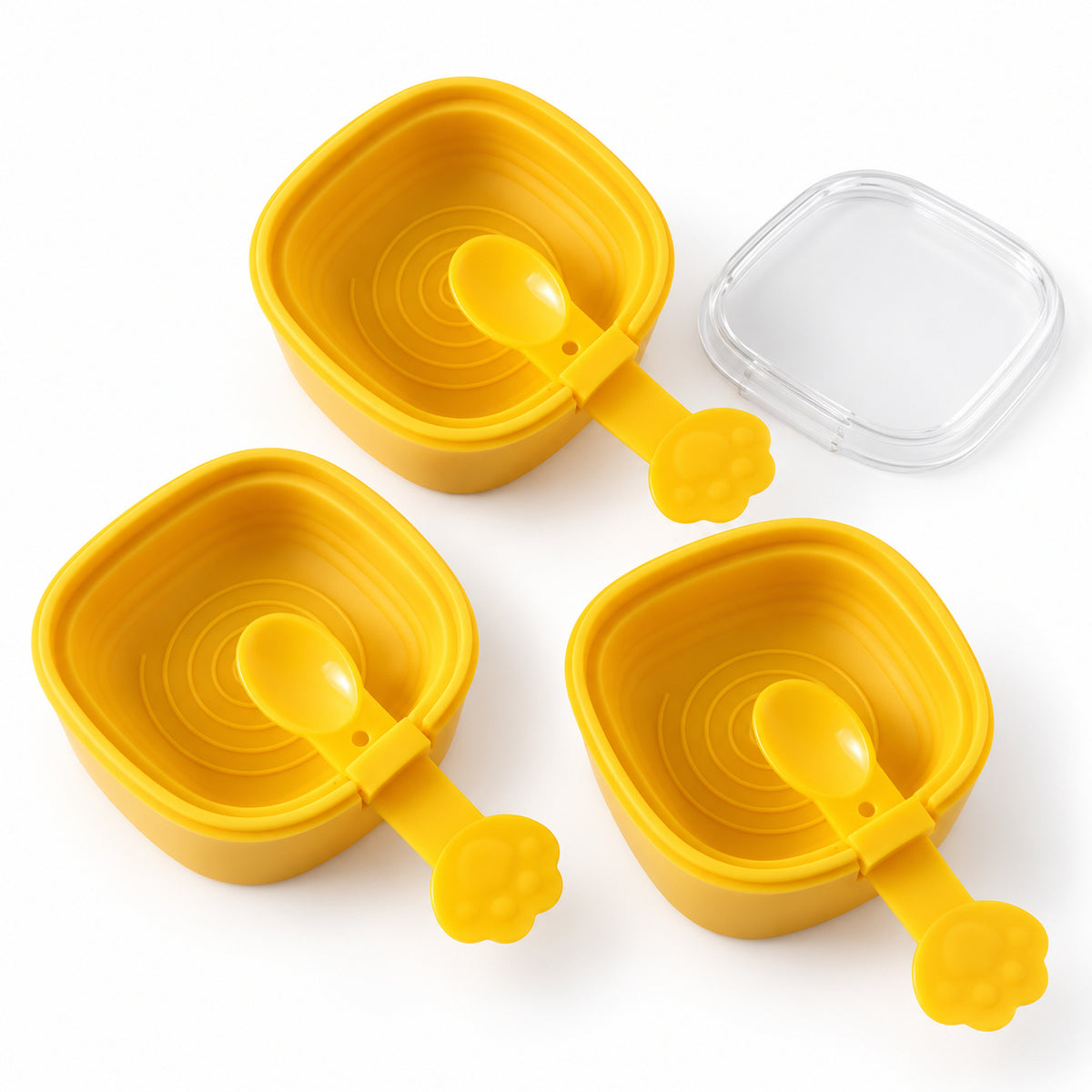 Three empty yellow square molds with attached paw-shaped spoons and a detached clear lid on a white surface.