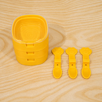A stack of three yellow square molds with a clear lid, and three separate yellow paw-shaped spoons on a wooden table.