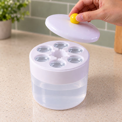 A hand lifts the white lid with a yellow knob from the ice cube maker, revealing six circular cavities filled with water.