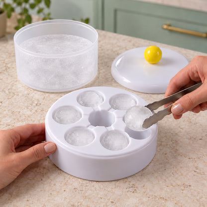 Hands use tongs to remove a round ice cube from the white tray, with other ice cubes in the tray and a container of ice