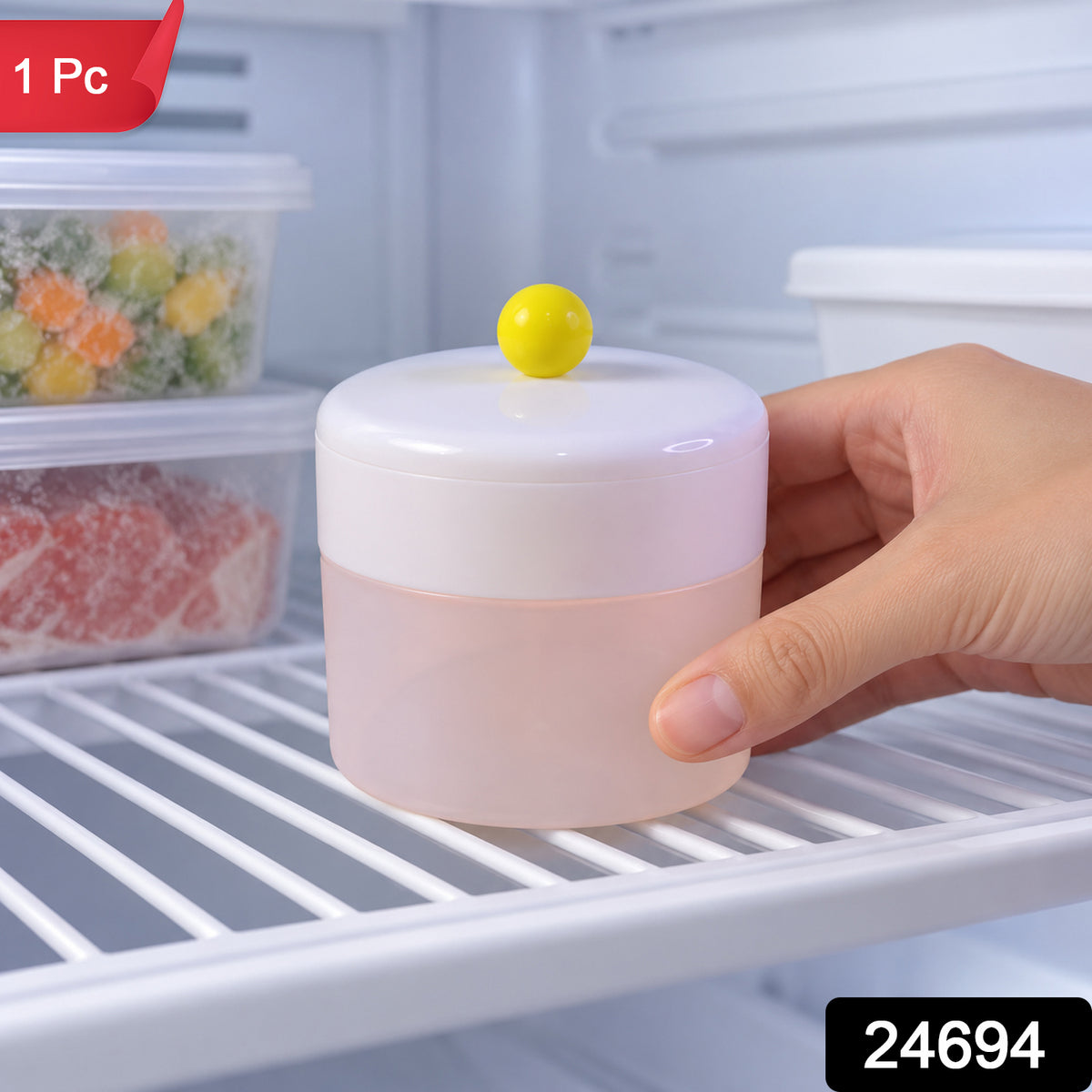 A hand places the white and pink round ice cube maker with a yellow knob on a refrigerator shelf, with frozen items in the