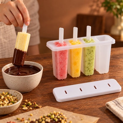 A hand dips a yellow popsicle into a bowl of melted chocolate, with a white 4-cavity mold holding three colorful popsicles.