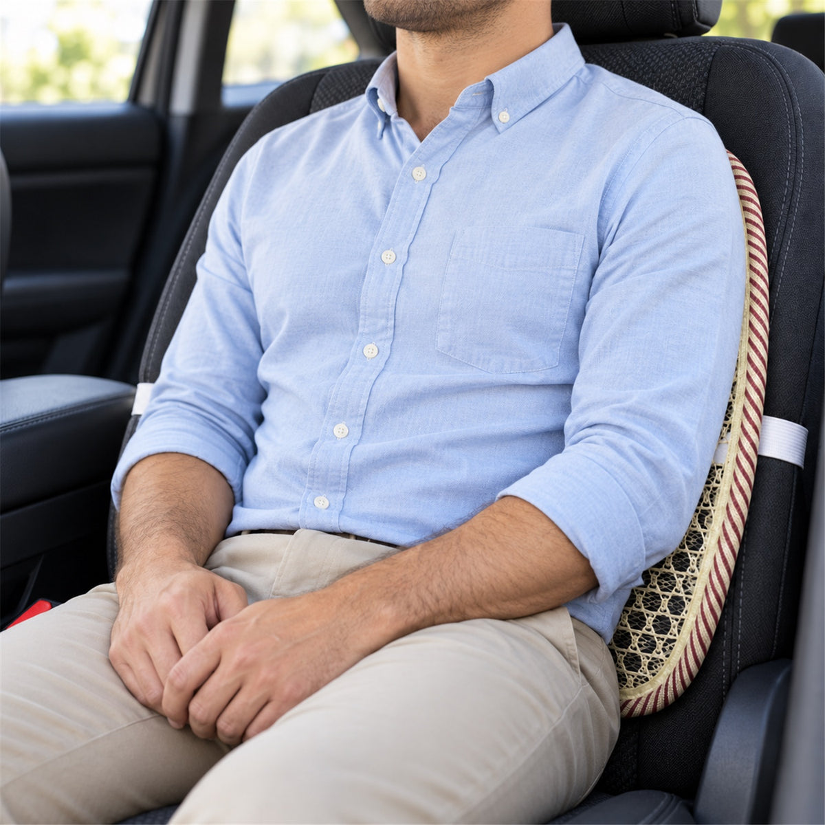 A man wearing a light blue shirt and khaki pants sits in a car, using a beige mesh back support cushion on a dark fabric