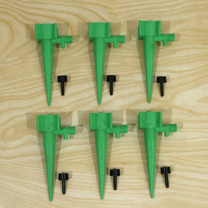 Six green plastic plant watering spikes and six small black caps are arranged on a light wooden surface.