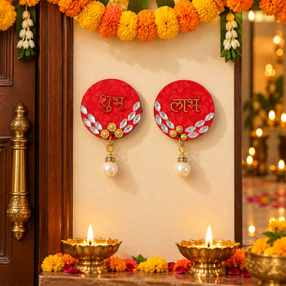 Two red circular Shubh Labh decorations with pearl hangings are affixed to a wall between two wooden doors, with lit diyas