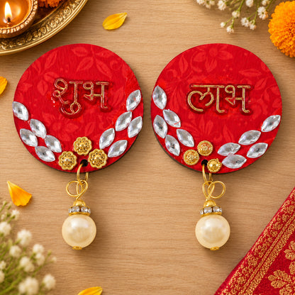 A pair of red circular Shubh Labh decor pieces with gold details and pearl drops rest on a light wooden surface next to a