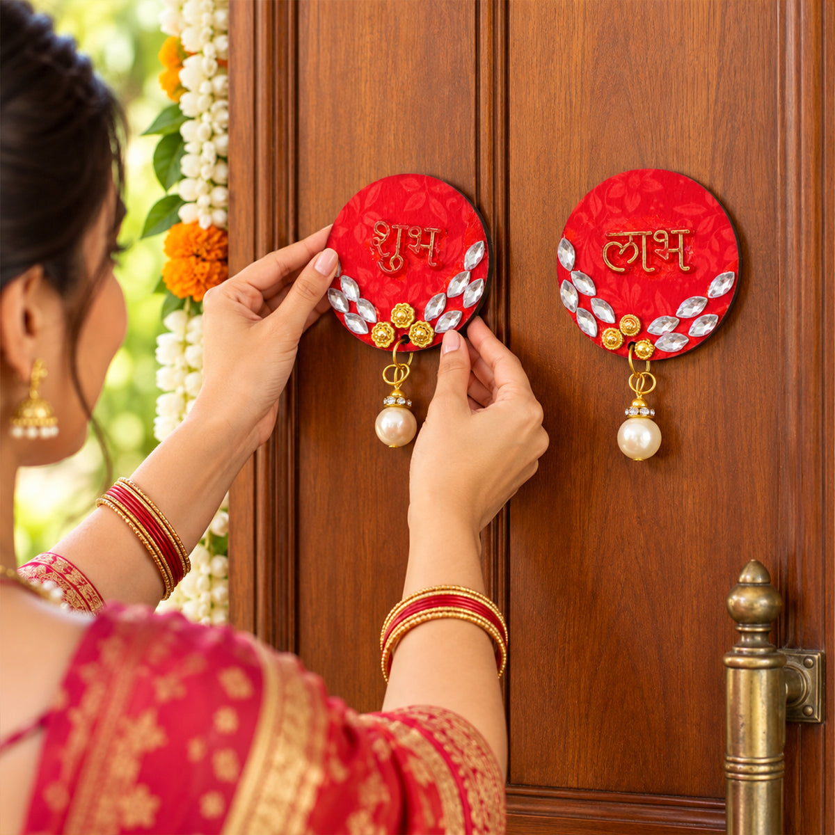 A person in traditional attire applies a red circular Shubh decoration with gold script and a pearl drop to a wooden door.
