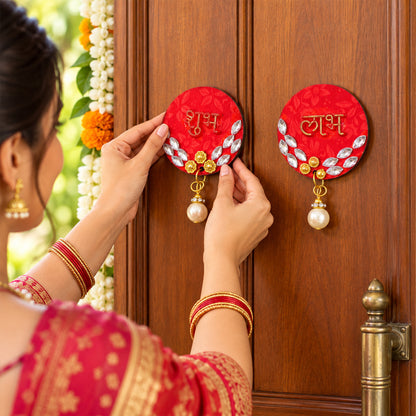 A person in traditional attire applies a red circular Shubh decoration with gold script and a pearl drop to a wooden door.
