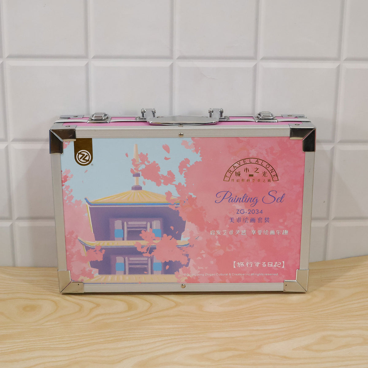 A closed pink metal art case with a pagoda and cherry blossom design rests on a light wooden surface.