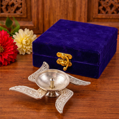 A silver Swastik design diya with a wick holder sits on a wooden surface next to a closed blue velvet box.