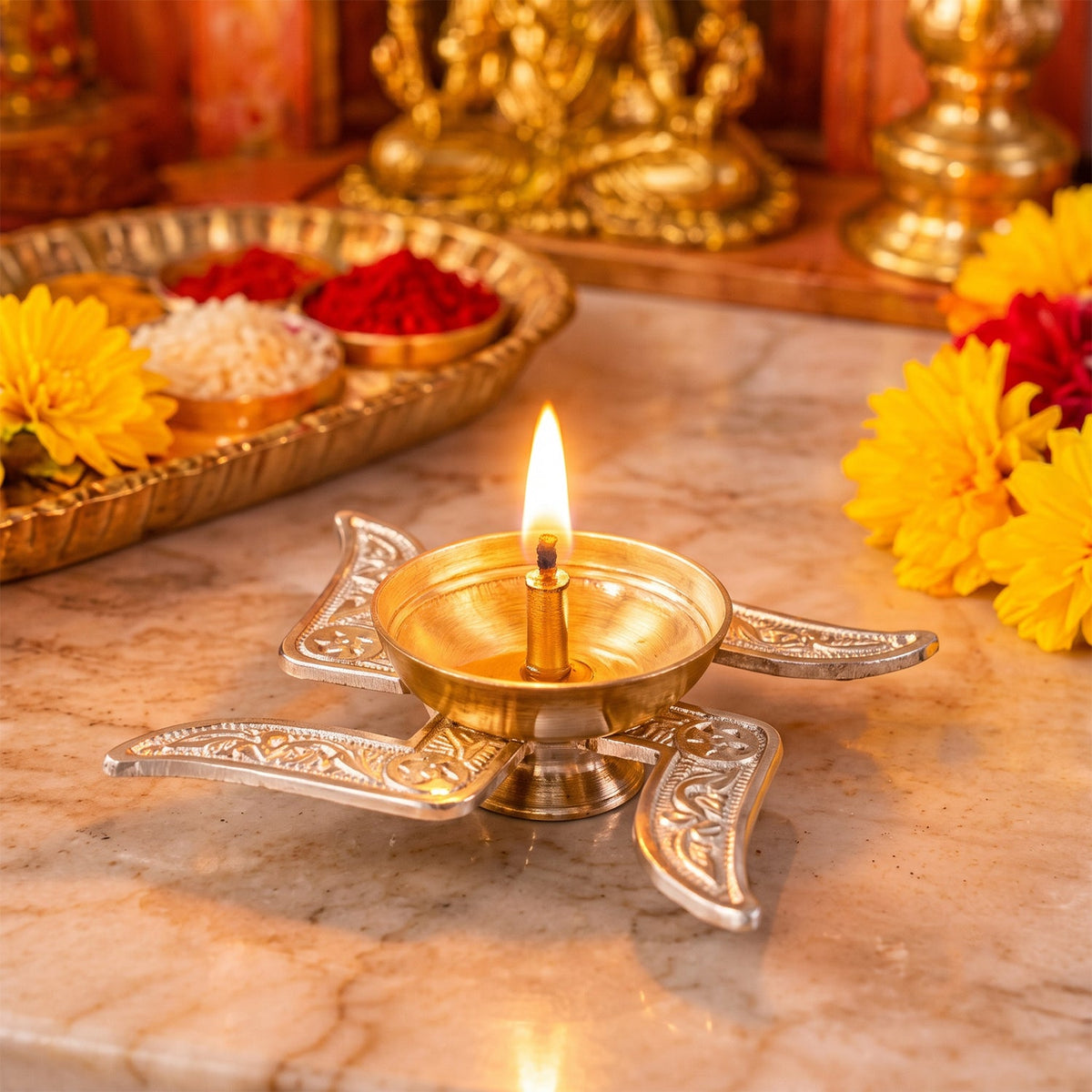 A lit silver Swastik diya with a golden bowl and a bright flame sits on a marble surface near a golden tray.