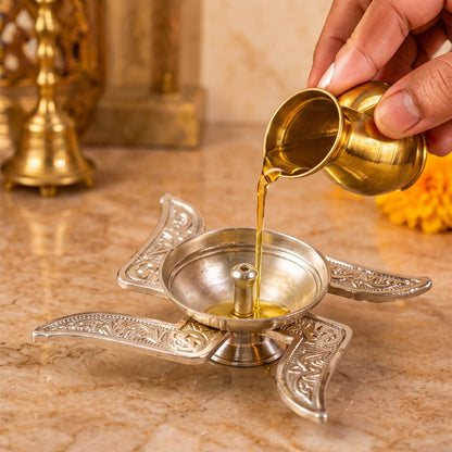 A hand pours oil from a small golden pot into the silver Swastik diya on a marble surface.