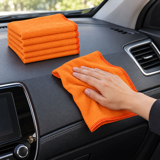 A hand wipes a car dashboard with an orange microfiber cloth, while a stack of five folded cloths sits nearby.