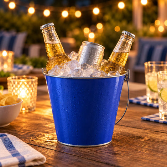 A blue metal bucket filled with ice, two beer bottles, and one soda can sits on a wooden table outdoors at night, surrounded